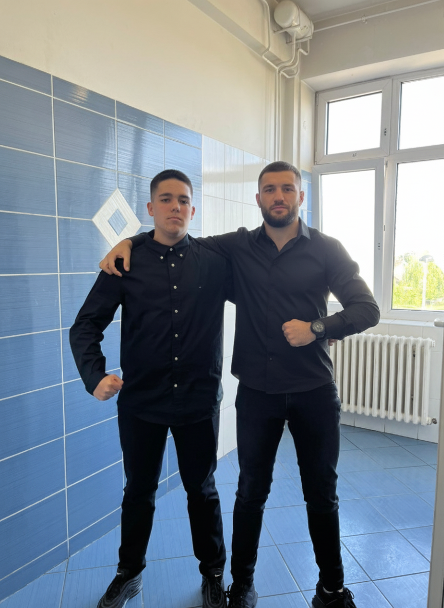 Two men are standing next to each other in a room with blue and white tiled walls. They are both wearing black shirts and black pants. The man on the left has his arm around the other man's shoulder.