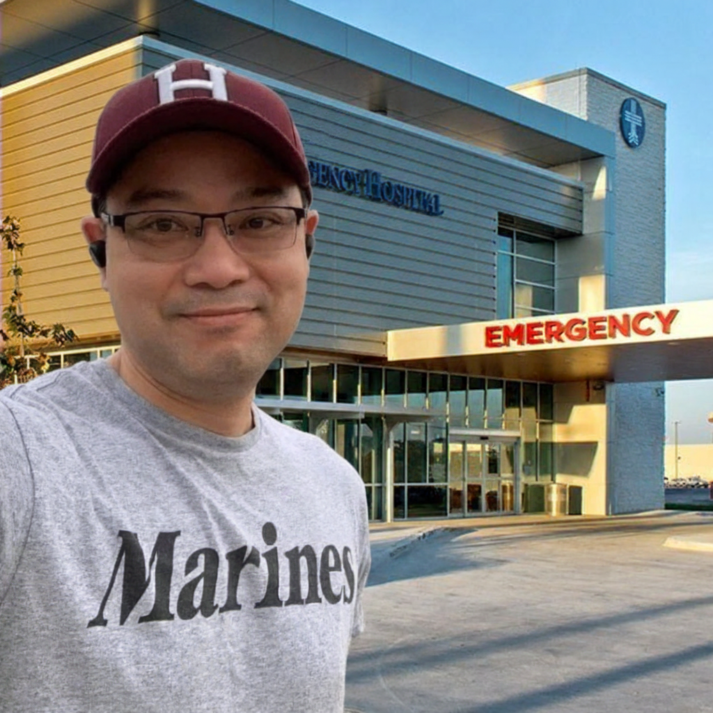 A man wearing a hat and a t-shirt with the word "Marines" on it is standing in front of a hospital emergency entrance.