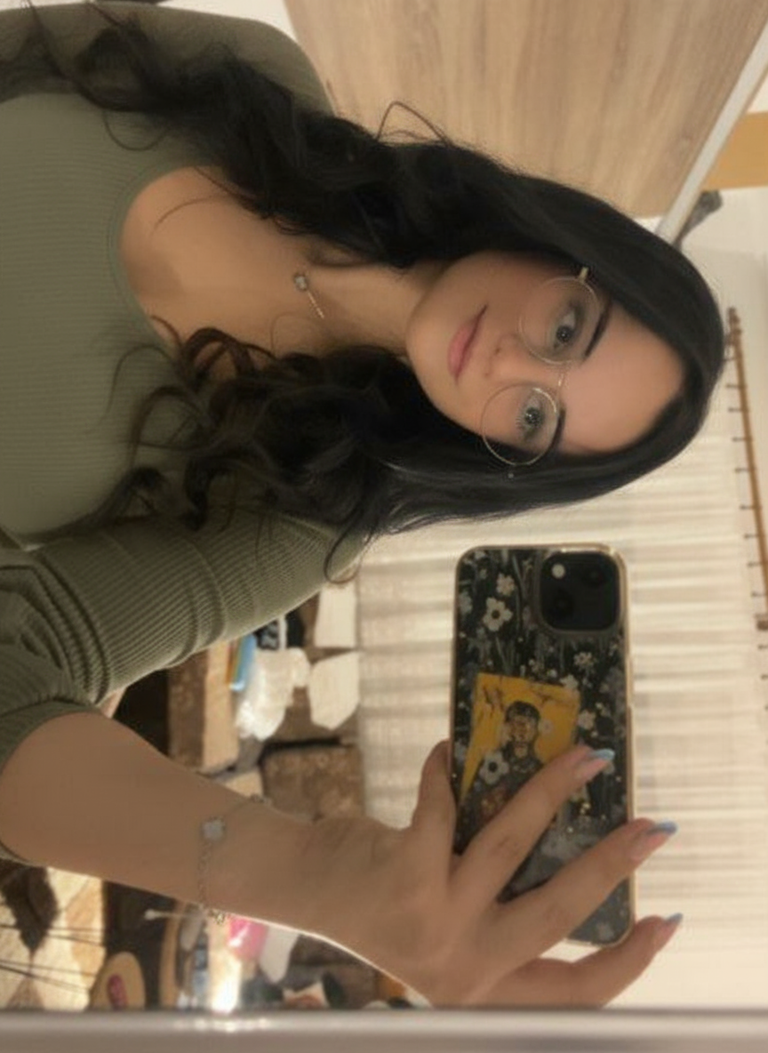 A woman with long dark hair and glasses is taking a selfie in a mirror. She is wearing a green top.