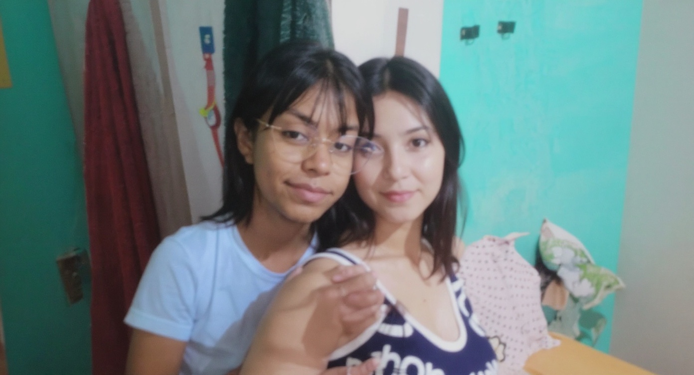Two young women are posing for a photo indoors. One is wearing glasses and a light blue t-shirt, while the other is wearing a tank top. They are standing close together.